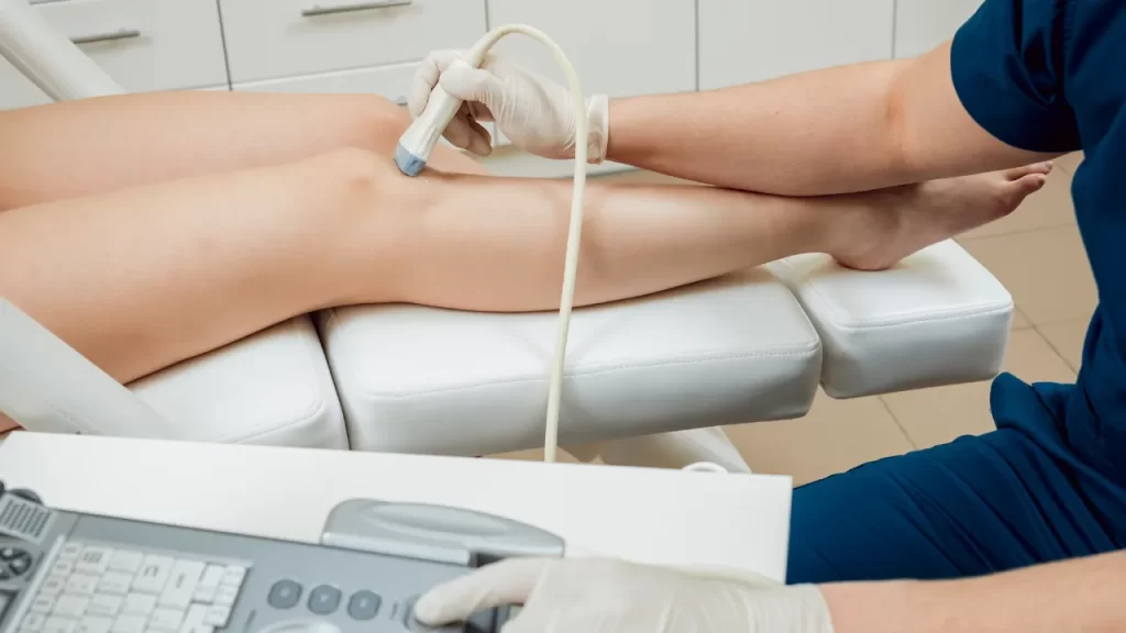Profissional de saúde realizando ultrassonografia vascular nas pernas de um paciente com o uso de transdutor e gel condutor.